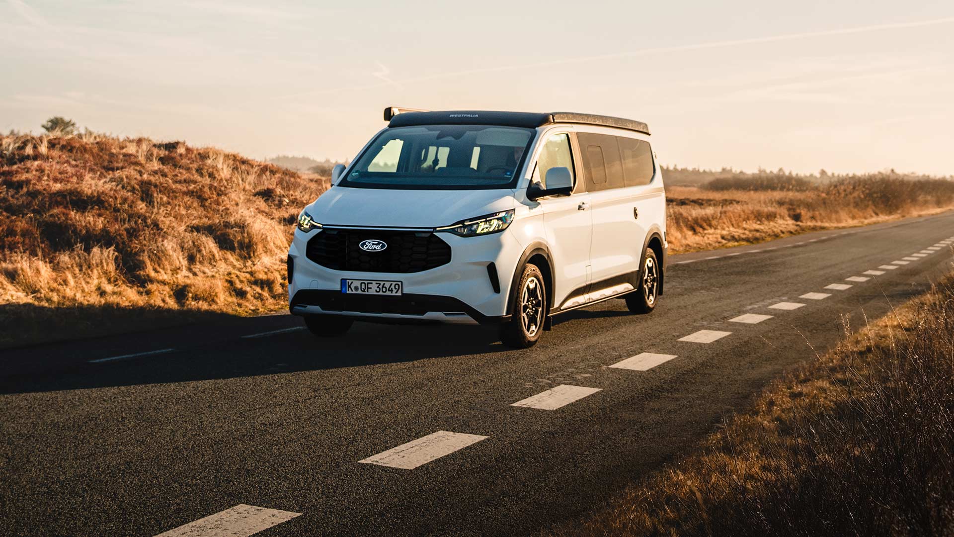 Frontansicht der Ford Nugget Jubiläumsedition während der Fahrt auf einer asphaltierten Landstraße durch eine Dünenlandschaft bei sonnigem Wetter.