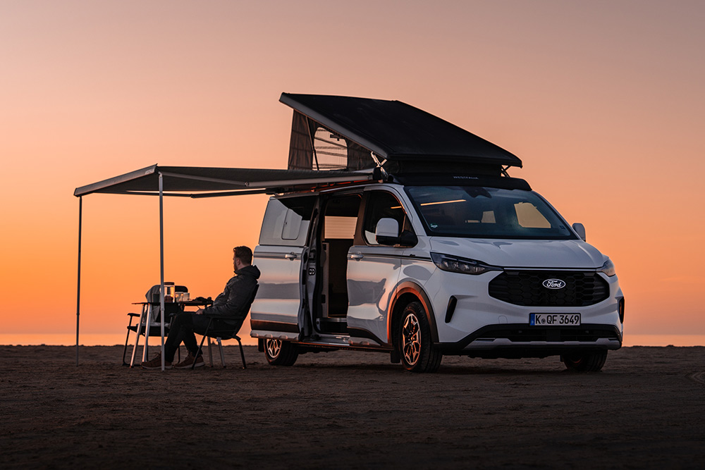 Ford Nugget Jubiläumsedition mit aufgestelltem Dach und ausgefahrener Markise in einer weitläufigen Landschaft bei Abenddämmerung. Eine Person sitzt im Campingstuhl unter der Markise.