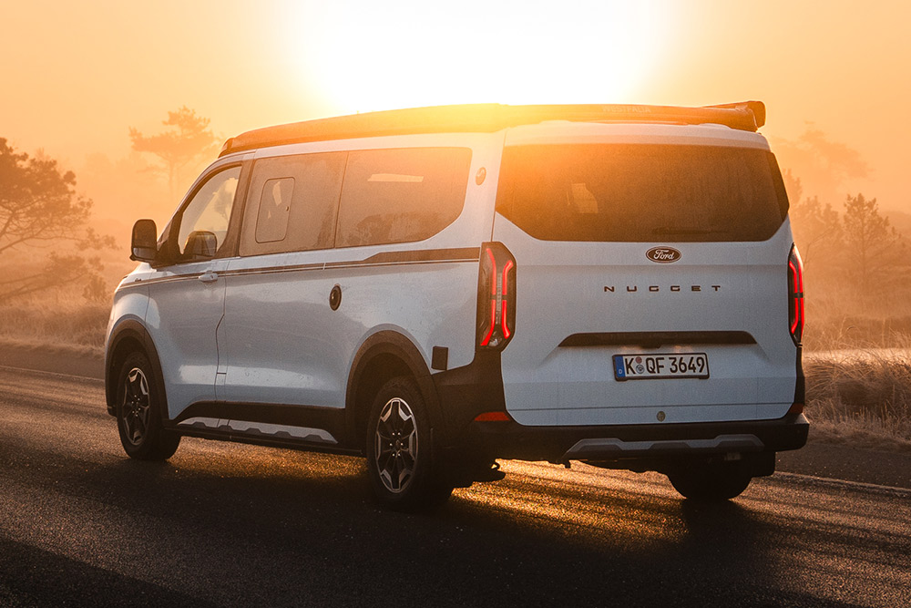 Heckansicht der Ford Nugget Jubiläumsedition auf einer asphaltierten Straße in einer nebligen Landschaft bei Sonnenaufgang.