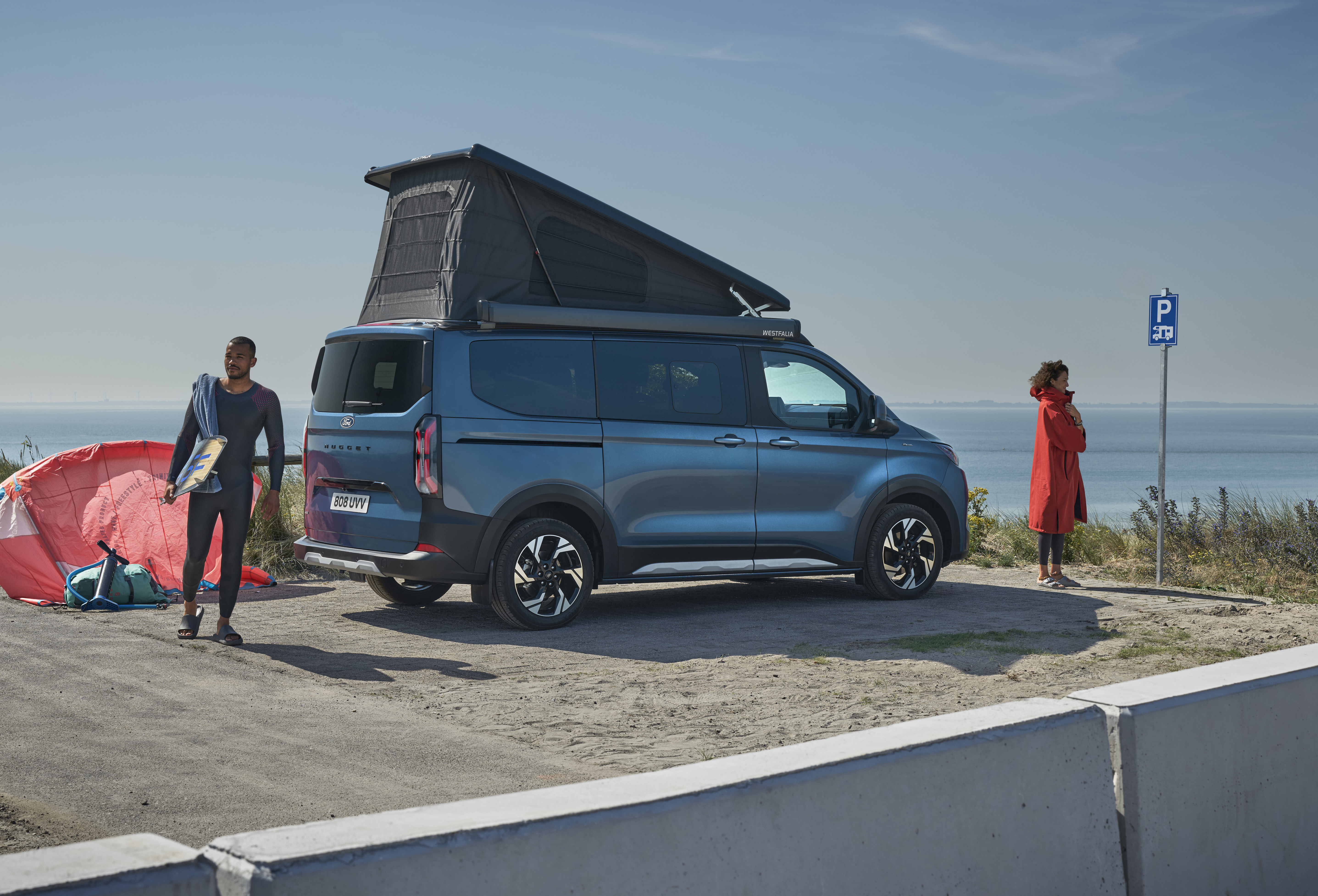 Ein dunkelblauer Ford Nugget mit aufgestelltem Dach parkt am Meer, während zwei Personen Kitesurf-Ausrüstung bereitlegen.