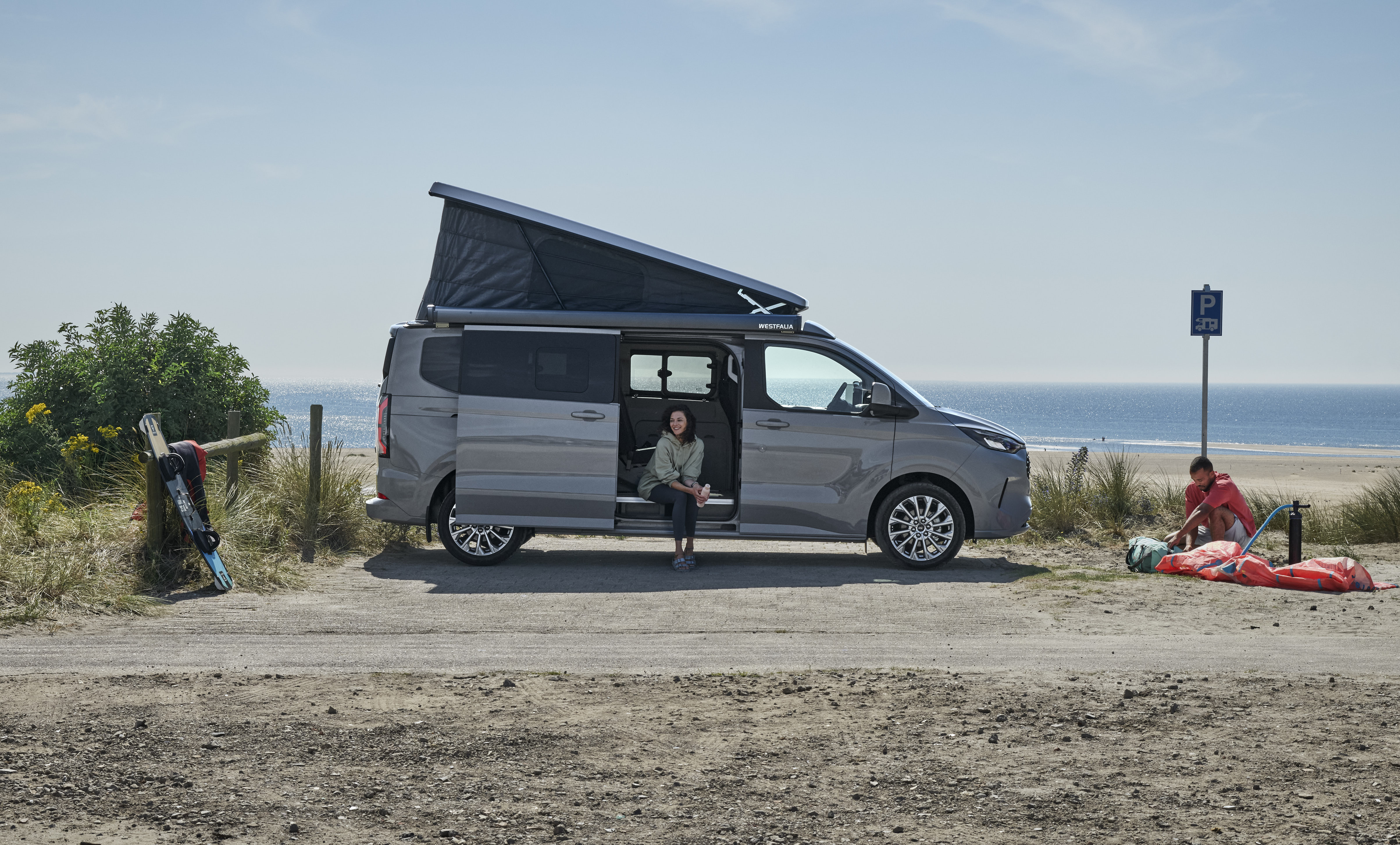 Ein grauer Ford Nugget mit aufgestelltem Dach steht am Strand, eine Person sitzt in der geöffneten Schiebetür.