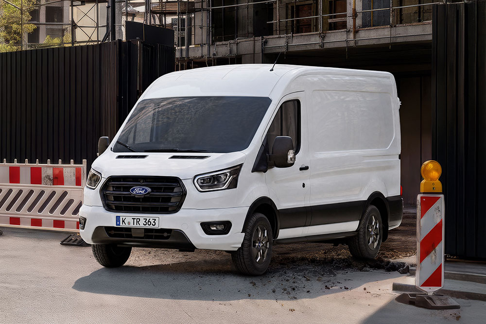 Ein weißer Ford Transit Kastenwagen steht auf einer Straße neben Baustellenabsperrungen und einer Warnleuchte. 