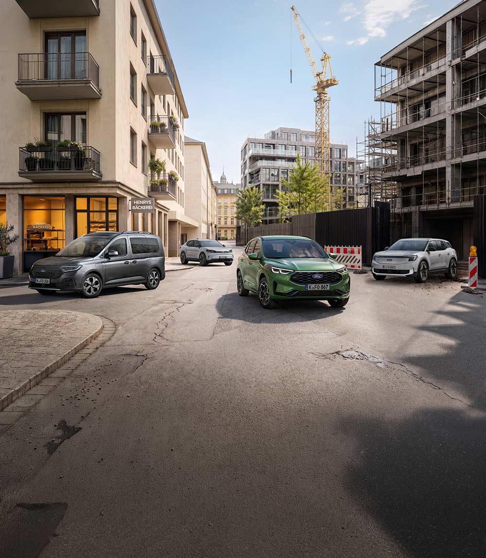 Ein grauer Tourneo Connect, ein grüner Kuga, ein silberner Capri und ein weißer Explorer stehen auf einer Stadtstraße.