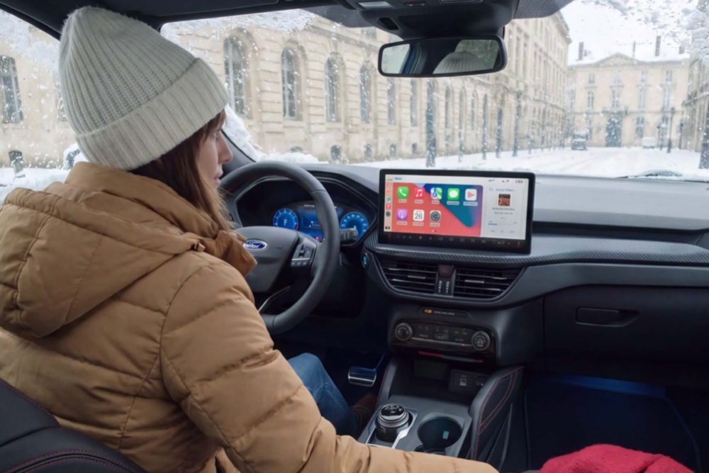 Eine Person in Winterkleidung sitzt im Ford auf dem Fahrersitz, ein Touchscreen zeigt eine Navigations-App.