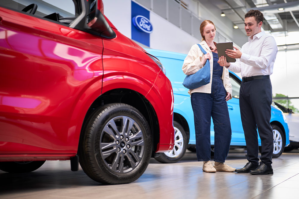 Zwei Personen in einem Ford-Showroom betrachten ein Tablet, mit einem roten Ford-Transporter im Vordergrund und einem blauen Ford-Transporter dahinter.