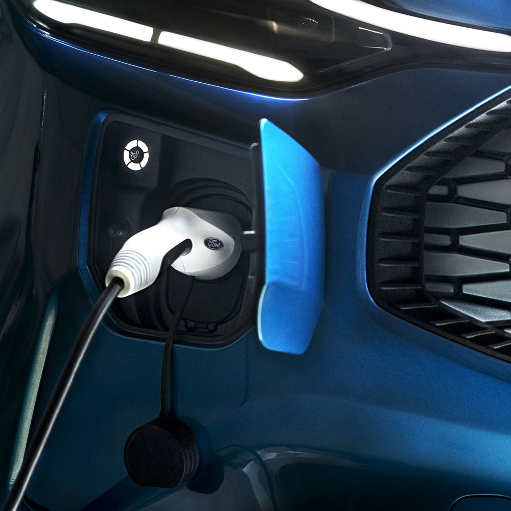 Eine Ladestation für Elektroautos mit angeschlossenem Ladegerät für Elektrofahrzeuge vor einem blauen Ford-Fahrzeug.