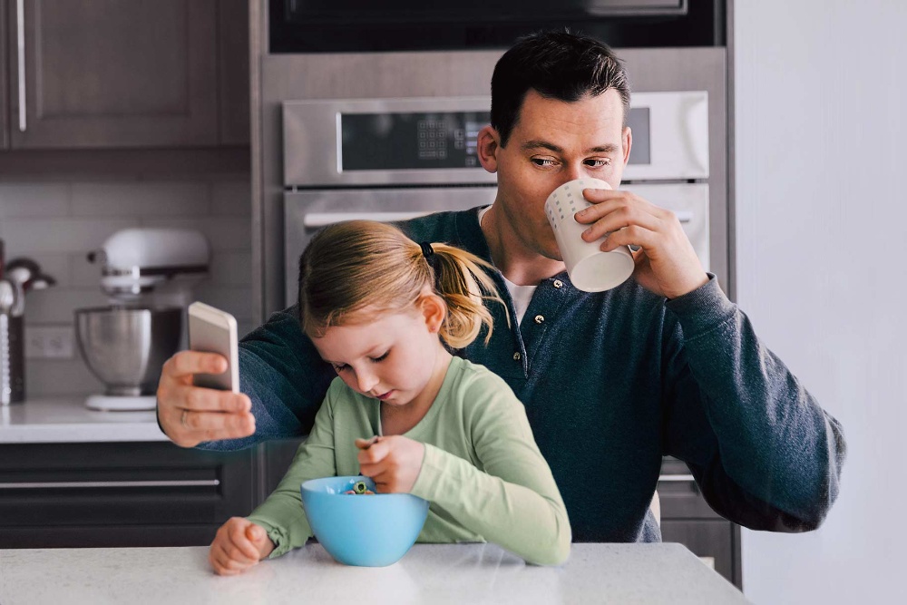 Mann mit Kind auf dem Schoss trinkt aus einer Tasse und schaut auf sein Smartphone