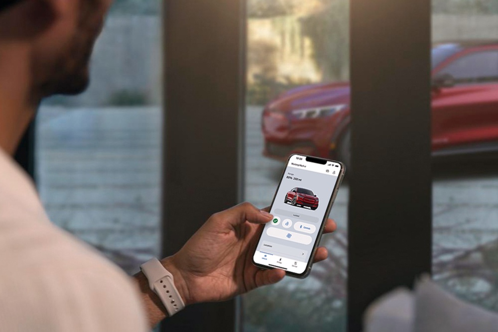 Ein Mann in seinem Wohnzimmer nutzt die Fahrzeug-Fernsteuerungsfunktionen in der FordPass-App für seinen in der Einfahrt geparkten Ford Mustang Mach-E