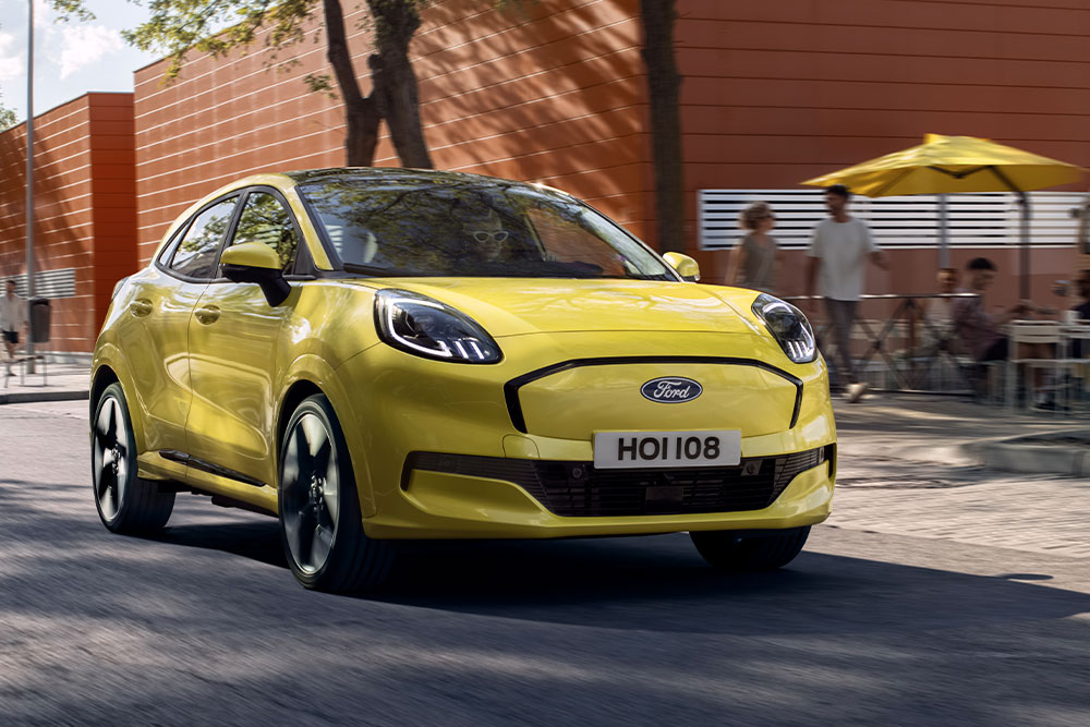 Frontansicht eines gelben elektrischen Ford Puma Gen-E®, der entlang einer Stadtstraße fährt.