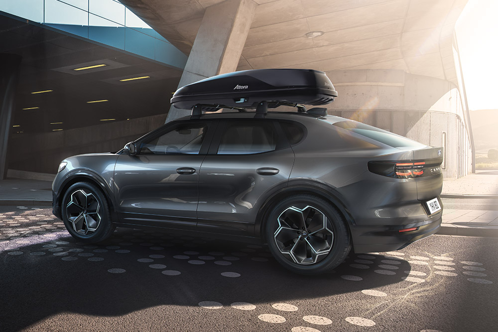 Seitliche 3/4-Ansicht eines roten Ford Capri mit Dachbox, geparkt unter einer modernen Gebäudestruktur.