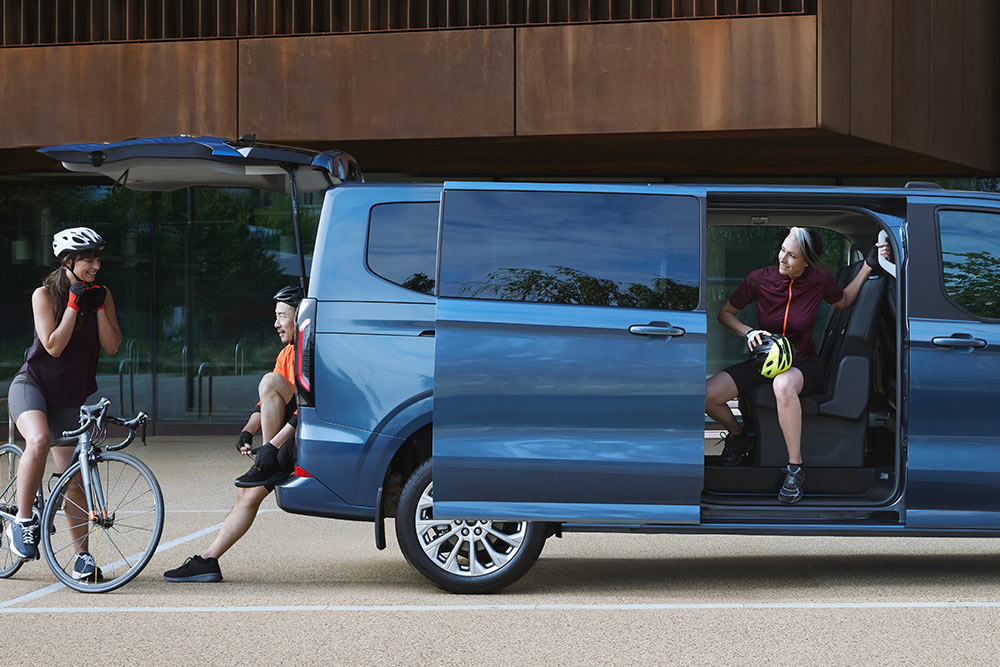 Blauer Ford Tourneo Custom parkt mit geöffneten Schiebetueren, Personen mit Fahrradern in der Naehe.
