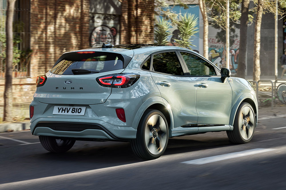 Rückansicht eines grauen Ford Puma, der auf einer Stadtstraße mit Menschen und Gebäuden fährt.