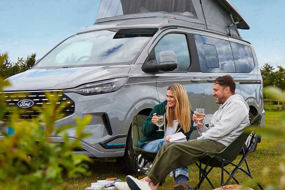 Ein Paar genießt Getränke in Campingstühlen neben einem grauen Ford Transit Custom Camper Van mit Aufstelldach.