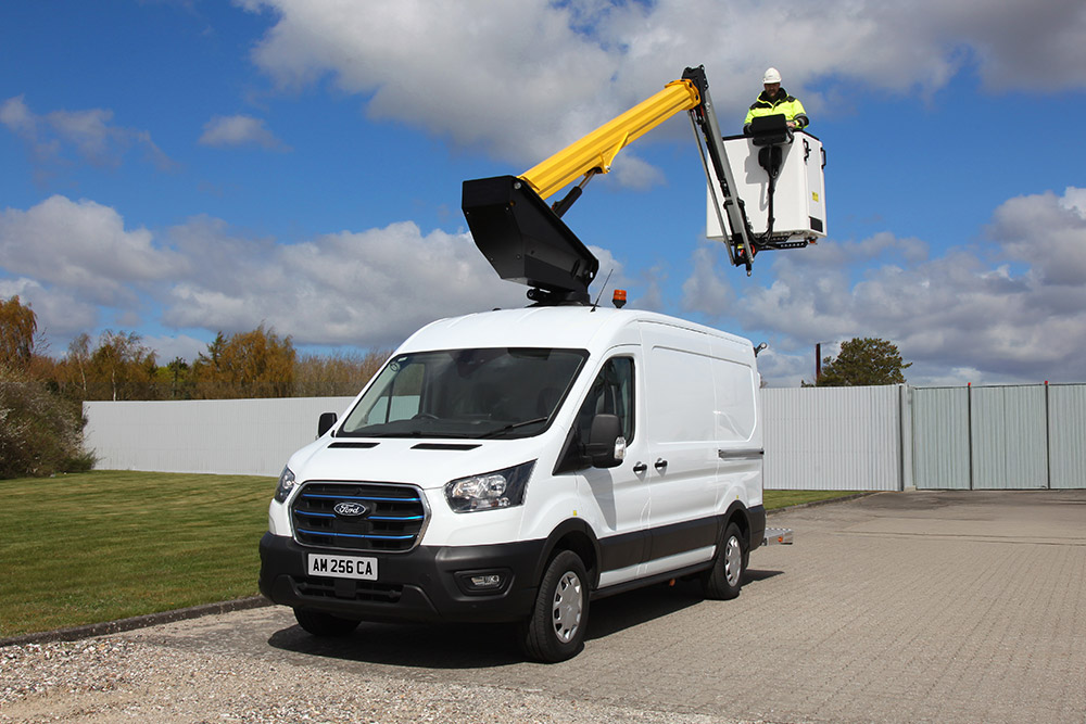 Ein weißer Ford Transit Transporter mit ausgefahrener Hebebühne, ein Techniker im Korb, unter blauem Himmel.