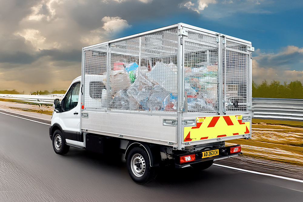 Rückansicht eines weißen Ford Transit Käfigkippers, voll mit Müll, fährt auf einer Straße.