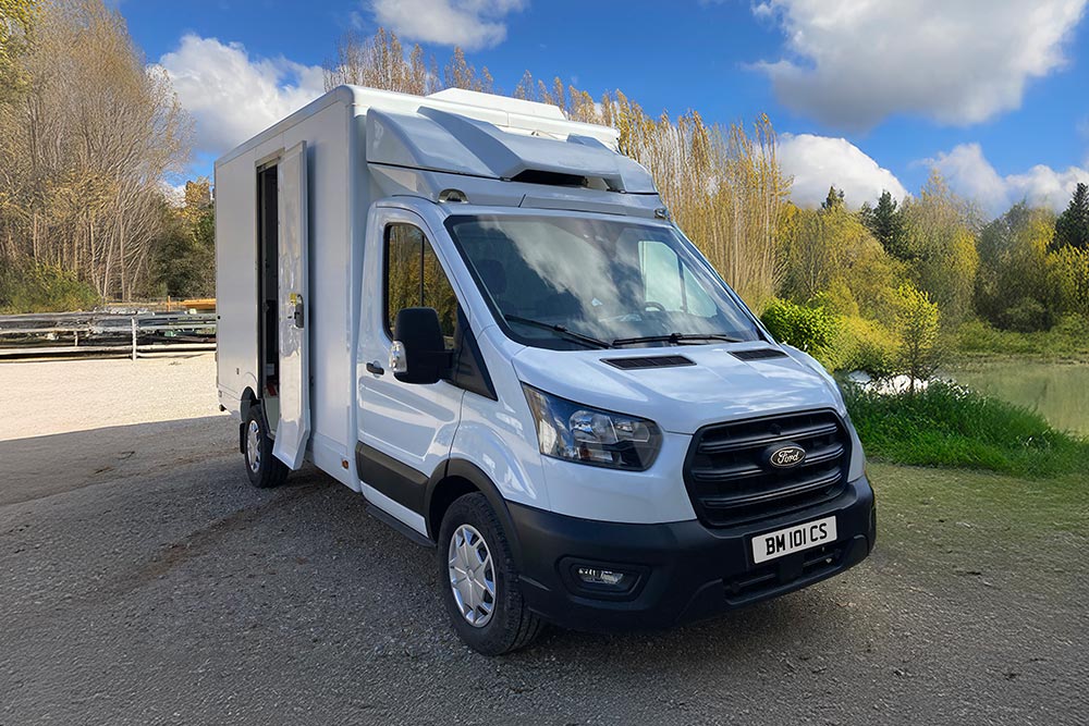 Vorderansicht eines weißen Ford Transit Kastenwagens, der an einem See mit Bäumen parkt.