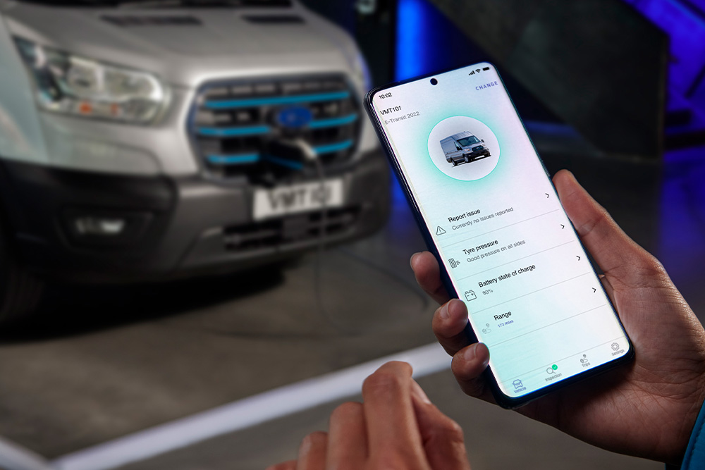 Eine Person hält ein Smartphone, das eine App-Oberfläche anzeigt, mit einem silbernen Ford Pro Transit Transporter, der im Hintergrund verschwommen ist.