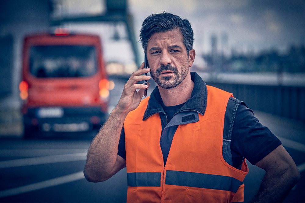 Arbeiter in orangefarbener Weste ruft Pannenhilfe auf einem Mobilgerät an.
