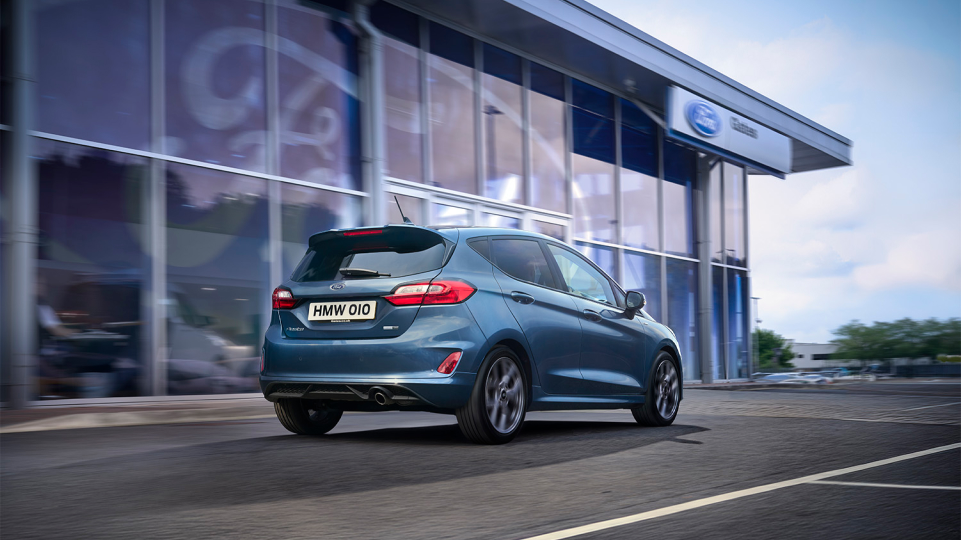 Blaues Ford-Fahrzeug fährt an einem Ford-Autohaus vorbei.